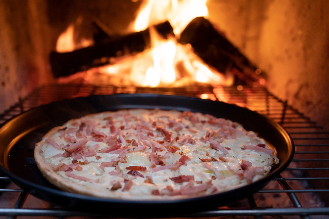 Wood-fired cheesy pizza topped with ham, cooking on a tray in front of an open flame