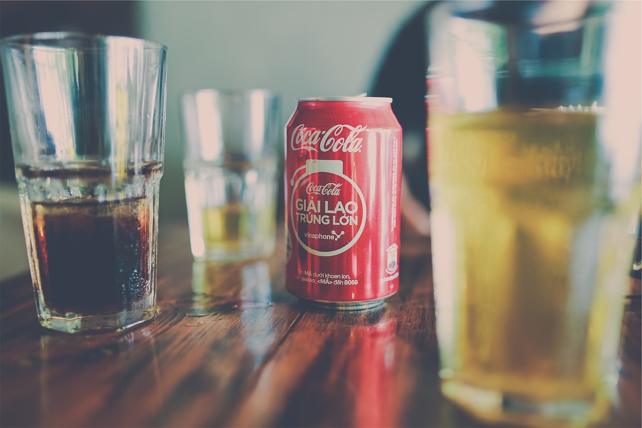 Red Coca-Cola can on a wooden table surrounded by glasses filled with various light and dark beverages.