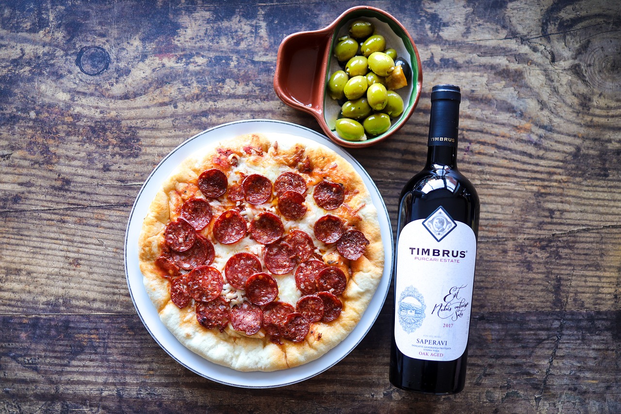 Pepperoni pizza on a white plate with a side of green olives in a small dish and a bottle of Timbrus red wine, set on a rustic wooden table