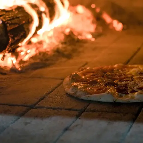 A pizza being cooked on the floor of a traditional oven, with wood burning and flames clearly visible in the background